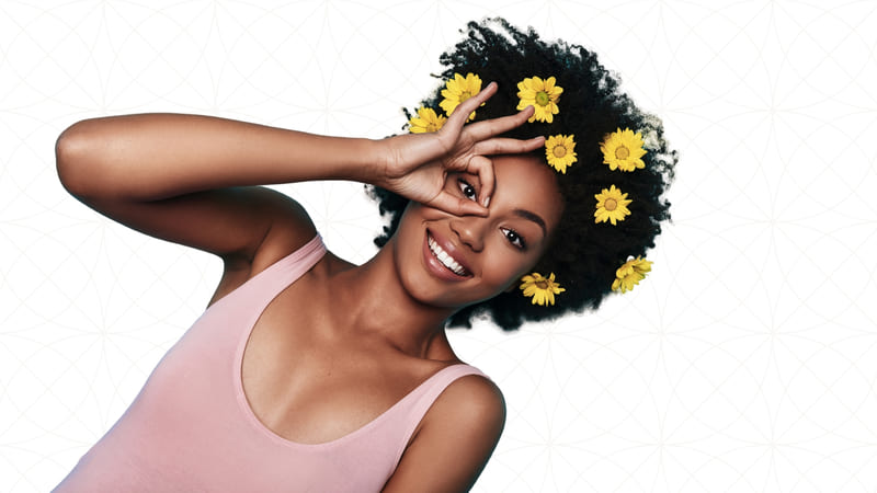 A young african american woman wearing eight yellow flowers on her hair as she makes a circle with her index and thumb and places is it around her eye as she looks through the middle