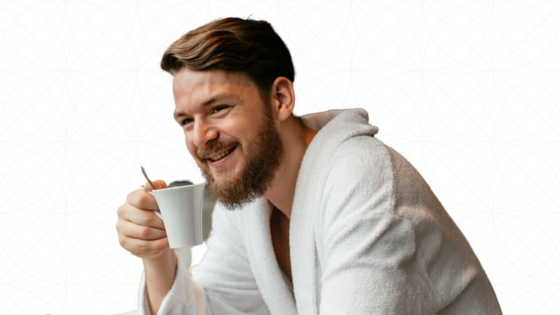 A young man with short hair, a beard and in a robe holding a cup of coffee as he sips on it