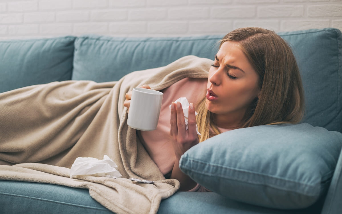 A young woman coughing into a tissues as she lays on her couch holding a coup of warm tea