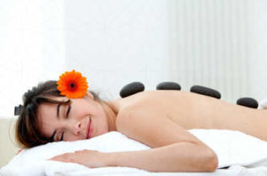 A young woman wearing an orange flower as she lays on a massage table with stones aligned in her back while she relaxes and smiles with her eyes closed