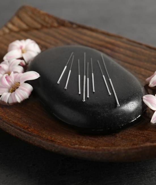 A set of acupuncture needles laying on a black stone surrounded by flowers on a wooden container